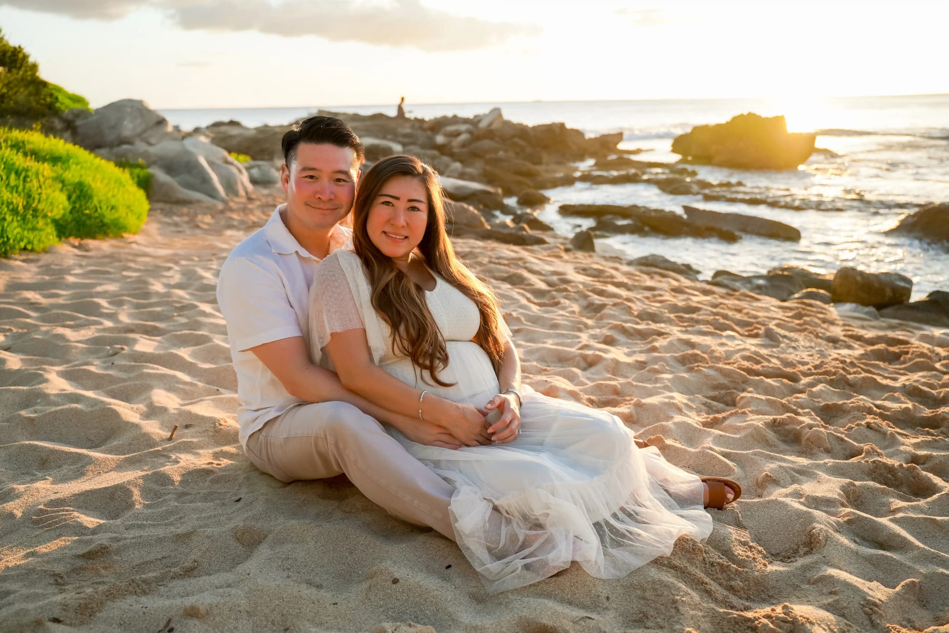 Maternity couple sitting on beach sand at golden hour